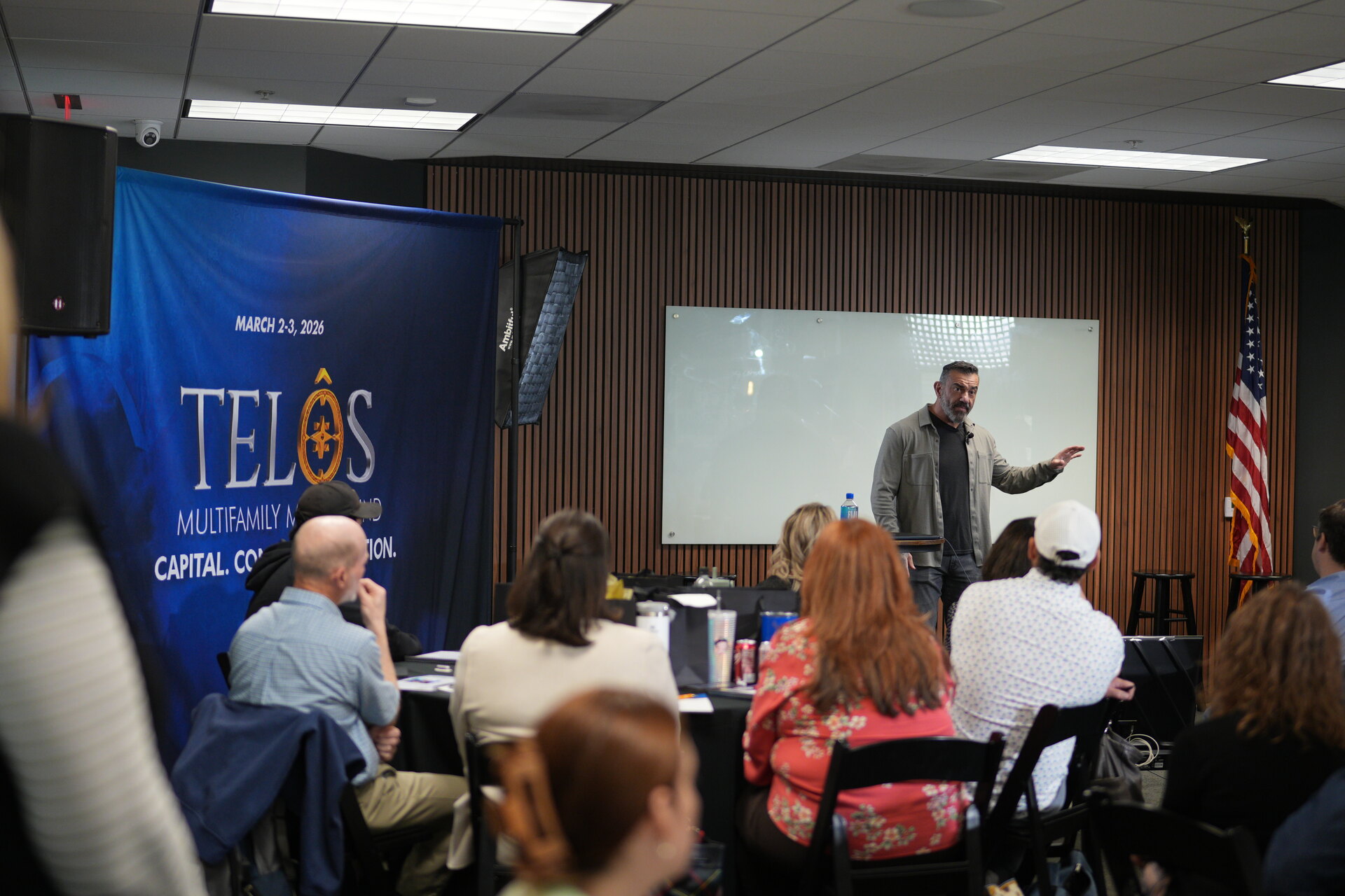 A speaker leading a March TELŌS session beside a whiteboard, with attendees seated close by